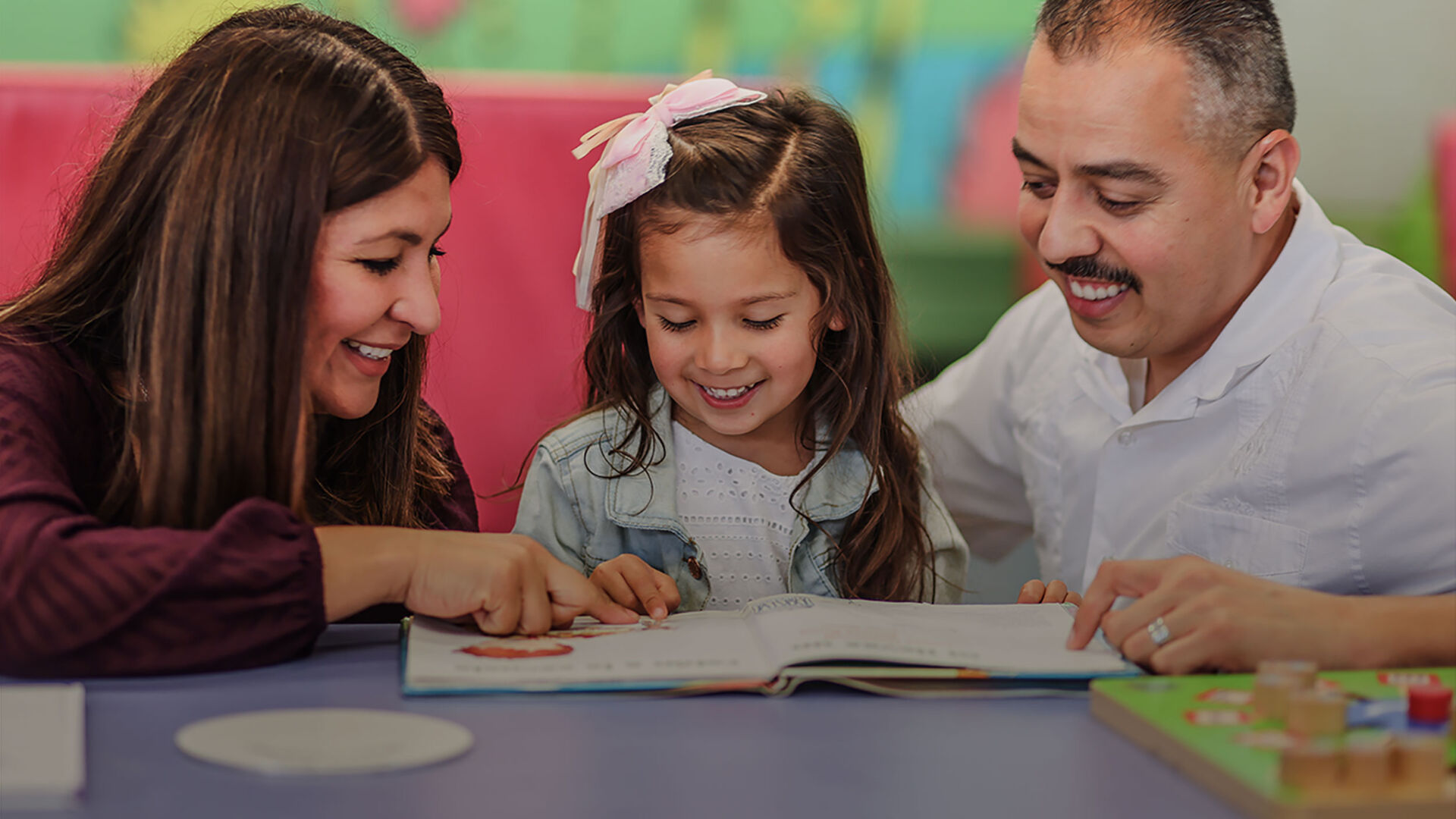 Photo of a smiling child reading a book with her parents